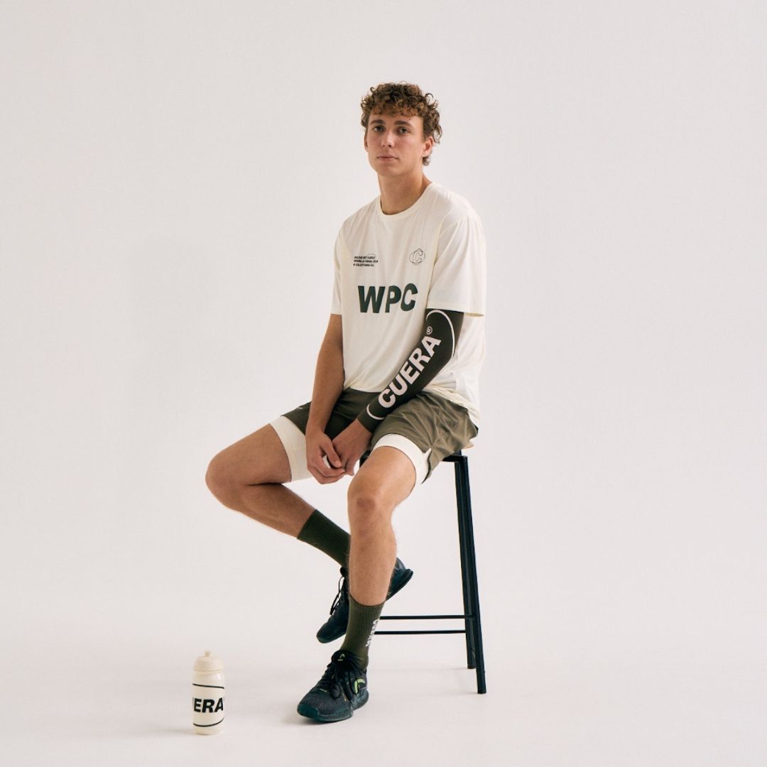 Model wearing Cuera Active WPC Light Weight T-Shirt in Off White / Army, lifestyle action shot on the padel court